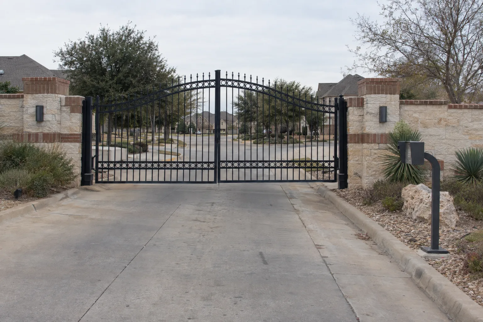 Ornamental iron swing gates at a gated HOA community entrance with brick columns, a keypad pedestal to the right, and suburban North Texas landscaping in warm late-afternoon light